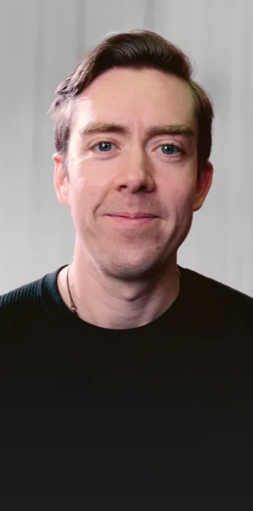 Portrait of a smiling man with short brown hair, light skin, and blue eyes wearing a black shirt in front of a light grey background.
